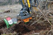 Das Bild zeigt die Maschine am Arm des Harvesters, die gerade ein Loch in die Erde gräbt. Man sieht auch ein grünes Gerät, das daran montiert ist mit zwei weißen Behältern in denen sich die Samen befinden.