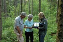 Zwei grauhaarige Menschen stehen mit einer jüngeren Frau im Wald. Die jüngere Frau ist in der Uniform einer Försterin gekleidet und zeigt den anderen beiden Menschen etwas auf einem Laptop