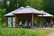 Hölzener Info- und Pausenpavillon am Waldrand, in welchem eine Familie gerade Brotzeit macht.