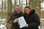 Ein Herr und eine Dame halten in einem winterlichem Wald eine Urkunde in die Kamera.