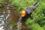 Mann mit Signalkappe liegt am Tümpel und schaut ins Wasser