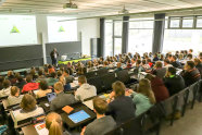 Studierende sitzen in einem Hörsaal und langweilen sich, während vorne ein Mann etwas vorstellt