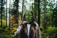 Ein echt hippes Boho-Girl, erkennbar an ihrer Kleidung, die auf den ersten Blick aus Naturmaterialien scheinen, hält sich dein Farnblatt vor das Gesicht, sodass nicht sie, sondern der Farn in der Kamera scharf ist. Der Wald hinter ihr verschwimmt im Bookeh.