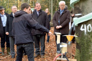 Das Bild zeigt im Vordergrund einen Schüler der eine an einem Baumstamm befestigte Apparatur erklärt. Zuhörer sind eine Gruppe Erwachsener, die im herbstlichen Wald stehen.