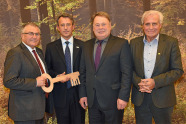 Vier Männer im Anzug stehen vor einem Waldbild. Die Zwei linksstehenden Männer halten einen großen Holzschlüssel