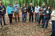 Menschen stehen im Halbkreis im Wald und haben sich mit roten Schnüren netzwerkartig verbunden