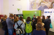 Schüler stehen vor einem Zentrum Wald Forst Holz Infostand und bekommen von einem Herrn etwas erklärt.