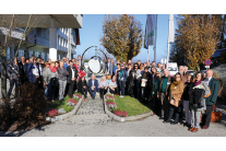 Eine große Gruppe Menschen steht im Sonnenschein auf einem Platz und guckt in die Kamera