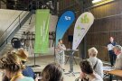 Eine Frau spricht am Mikro vor voll besetzter Maschinenhalle
