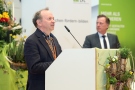 Einführungsvortrag von Dr. Hans Jürgen Reents. Er spricht. Im Hintergrund Prof. Dr. Kurt-Jürgen Hülsbergen, Blumengestecke und Rollups der HSWT und LfL.