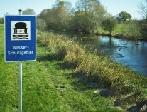 Ein Schild „Wasserschutzgebiet“ auf der Wiese neben einem Fluss.