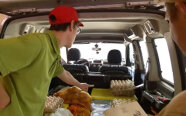 Ein junger Mann steht vor einem geöffneten Kofferraum und sortiert Waren.