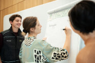Eine Frau schreibt auf ein Flipchart umrahmt von zwei weiteren Personen