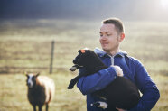 Ein junger Mann steht auf der Weide, hält ein kleines Lamm in seinem Arm und lächelt.