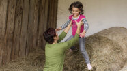 Eine Frau hebt ein lachendes Kind von großen Strohrundballen herunter.