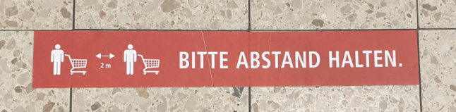 Auf einen Fliesenboden geklebte Markierung mit der Aufschrift "Bitte Abstand halten"