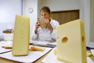 Eine Testerin im weißen Kittel sitzt am Tisch und riecht an einem größeren Eckstück Käse.