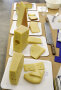 Fünf verschiedene Käsesorten sind in größere und kleinere Stücke geschnitten und auf einem Tisch auf eckigen Papptellern platziert.