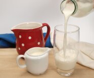 In ein Glas wird Milch aus einer Flasche geschüttet. Daneben steht ein kleines Sahnekännchen und ein roter Milchkrug mit weißen Tupfen. (Foto: LfL)