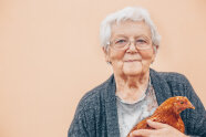 Eine ältere, grauhaarige Dame hält ein Huhn in Armen. Zu sehen sind Oberkörper und Kopf der Seniorin vor abricotfarbenem Hintergrund.